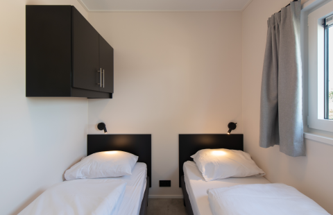 Schlichtes Lodge-Zimmer mit zwei Einzelbetten, dunklem Schrank, Leselampen und Fenster mit Vorhang.