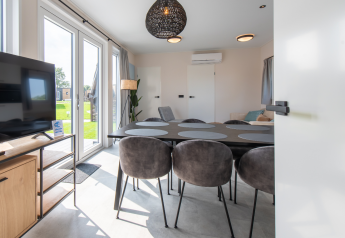 Lyst og moderne lodge interiør med spisebord, seks stole, tv og store vinduer med udsigt til haven.