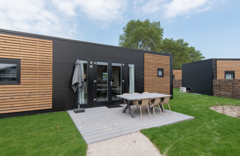 Lodge moderno con fachada en negro y madera, comedor exterior y césped verde bajo un cielo parcialmente nublado.