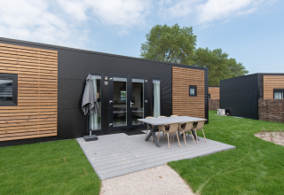 Lodge moderno con fachada en negro y madera, comedor exterior y césped verde bajo un cielo parcialmente nublado.