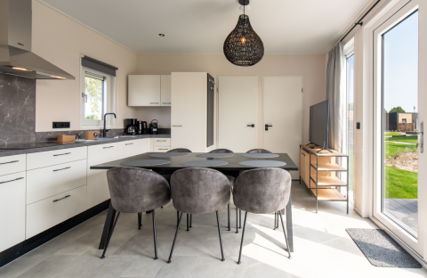 Modern lodge kitchen and dining area with grey chairs, large windows, and garden view in sunlight.