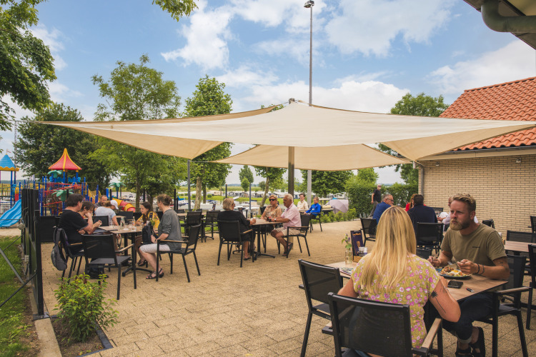 Außenterrasse von Villa Maritiem, Gäste essen unter Sonnensegeln neben einem Spielplatz an einem sonnigen Tag.