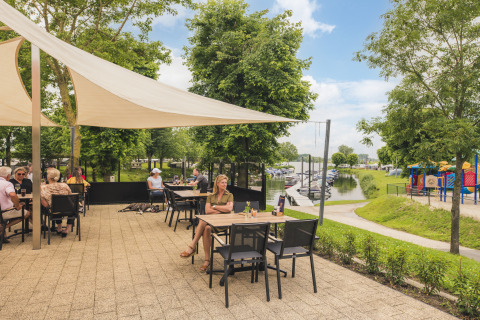 Udendørs terrasse på Villa Maritiem med gæster, udsigt over marinaen og legeplads på en solrig dag.