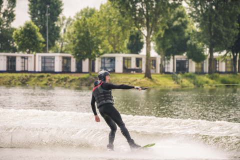 Persona practicando wakeboard en un lago frente al lodge Villa Maritiem, rodeado de árboles y cabañas.