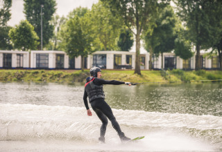 En person står på wakeboard på en sø foran Villa Maritiem lodge, omgivet af træer og hytter.