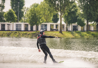 Persona practicando wakeboard en un lago frente al lodge Villa Maritiem, rodeado de árboles y cabañas.