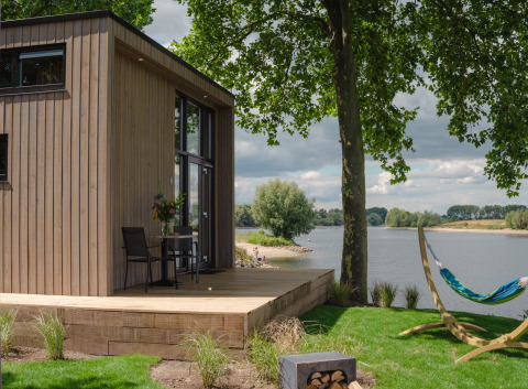 Moderne tiny house aan de rivier in Nederland met houten terras, hangmat en uitzicht op het water.