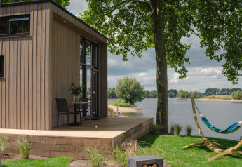 Modern tiny house by a Dutch riverside with wooden deck, hammock, scenic water view and lush greenery.