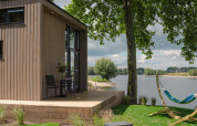 Moderne tiny house ved flodbredden i Holland med træterrasse, hængekøje og grøn udsigt til vandet.