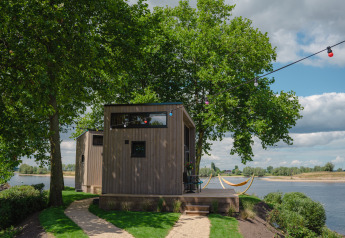 Casa pequeña de madera junto al río con hamacas, luces de colores y árboles en MarinaPark Bad Nederrijn.