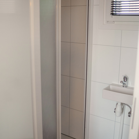 Lille badeværelse med hvidt flisegulv, et lille håndvask og vindue i Tiny River House i Holland.