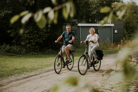 Dos personas mayores pasean en bicicleta por un camino de tierra junto a una lujosa tiny house en Camping Si-Es-An, Holanda.