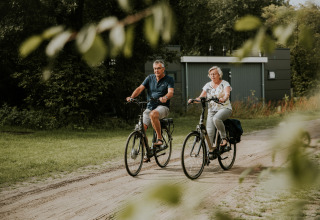 Dos personas mayores pasean en bicicleta por un camino de tierra junto a una lujosa tiny house en Camping Si-Es-An, Holanda.