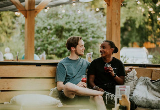 Dos personas sentadas en un banco exterior en un alojamiento glamping, compartiendo bebidas y sonrisas.