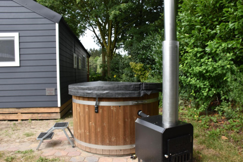Bañera de hidromasaje de madera con tapa negra y estufa junto a casa azul en Camping 't Veld, Países Bajos.