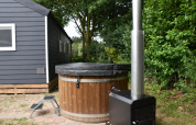 Vasca idromassaggio in legno con copertura nera e stufa accanto a uno chalet blu a Camping 't Veld.