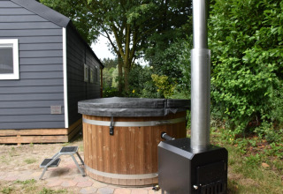 Holz-Whirlpool mit schwarzer Abdeckung und Ofen neben einem grauen Chalet im grünen Garten.