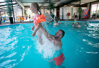 Mann spielt lachend mit Mädchen im Hallenbad im Beach House mit Whirlpool, Camping ’t Veld, Niederlande.