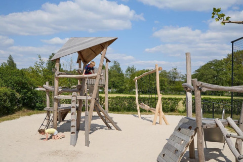 Children play on a wooden playground with sand, under a blue sky at Camping 't Veld in the Netherlands.
