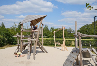 Niños juegan en un parque infantil de madera con arena bajo un cielo azul en Camping 't Veld, Países Bajos.
