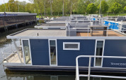 Husbåd navngivet Havenlodge ved Marina Parcs Naarden i Holland, fortøjet ved havnen med grøn omgivelse.