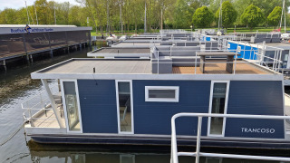 Casa flotante llamada Havenlodge en Marina Parcs Naarden, Países Bajos, amarrada en un puerto rodeado de árboles.