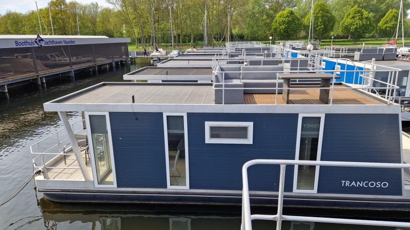 Casa flotante llamada Havenlodge en Marina Parcs Naarden, Países Bajos, amarrada en un puerto rodeado de árboles.