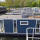 Casa flotante llamada Havenlodge en Marina Parcs Naarden, Países Bajos, amarrada en un puerto rodeado de árboles.