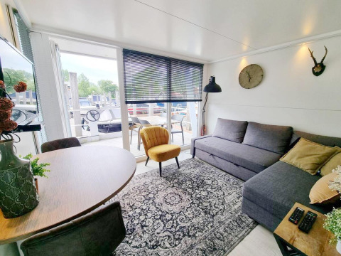 Sala luminosa en la casa flotante 'De Hoop' en Marina Parcs, Países Bajos, con sofá, sillón amarillo y vista al muelle.