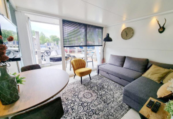 Lys stue på husbåd 'De Hoop' i Marina Parcs, Nederlandene, med sofa, gul stol og udsigt til dækket.