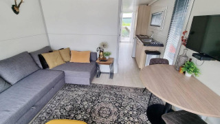 Moderne zithoek op woonboot 'De Hoop' bij Marina Parcs, met grijze zetel en open keukentje.