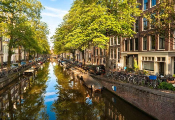 Vista desde la casa flotante De Hoop en Marina Parcs, Ámsterdam, con canal, botes, árboles y bicicletas.