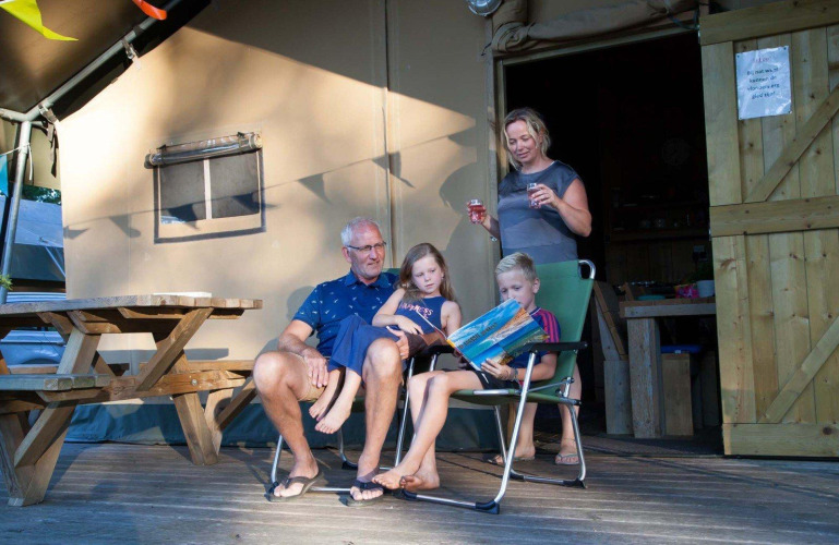 Familia relajándose fuera de una tienda safari en Recreation Park Den Blanken, Países Bajos, niños leyendo.