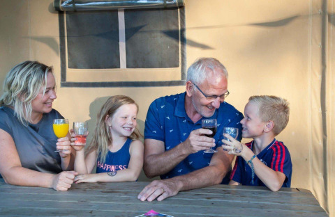 Familie stoßt mit Getränken an, sitzt vor einem Safarizelt im Recreation Park Den Blanken, Niederlande.