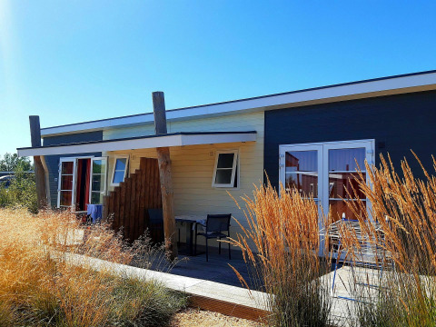 Cabaña Comfort Lodge en Kompas Camping - Westende, Bélgica, rodeada de hierbas altas y cielo azul brillante.