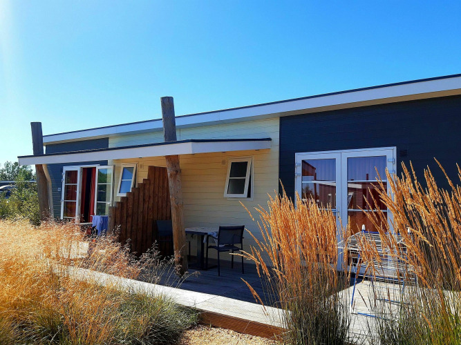 Cabaña Comfort Lodge en Kompas Camping - Westende, Bélgica, rodeada de hierbas altas y cielo azul brillante.