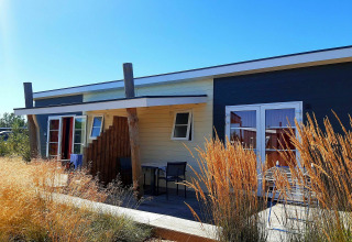 Cabaña Comfort Lodge en Kompas Camping - Westende, Bélgica, rodeada de hierbas altas y cielo azul brillante.