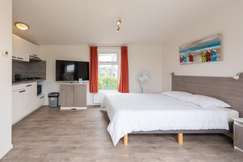 Binnenzicht van Comfort Lodge tiny house met keuken, tweepersoonsbed, tv, ventilator en rode gordijnen.