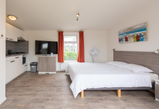 Binnenzicht van Comfort Lodge tiny house met keuken, tweepersoonsbed, tv, ventilator en rode gordijnen.