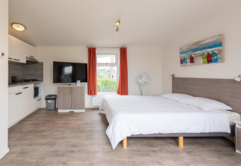 Binnenzicht van Comfort Lodge tiny house met keuken, tweepersoonsbed, tv, ventilator en rode gordijnen.
