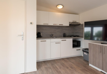 Cucinotto moderno e zona giorno nella tiny house Comfort Lodge con mobili bianchi, TV e pavimento in legno.