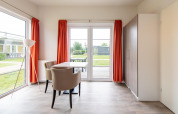 Lichte leefruimte in Comfort Lodge tiny house met grote ramen, oranje gordijnen en eettafel.