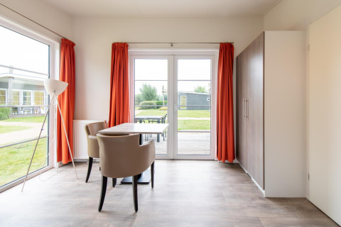 Lichte woonkamer in Comfort Lodge tiny house met grote ramen, oranje gordijnen en eettafel.