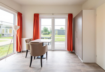 Sala luminosa en la tiny house Comfort Lodge con grandes ventanas, cortinas naranjas y mesa de comedor.