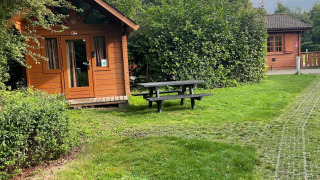 Foto van een Basic blokhut bij Urban-Gardens Ieper, België, met gras, struiken en een picknicktafel.