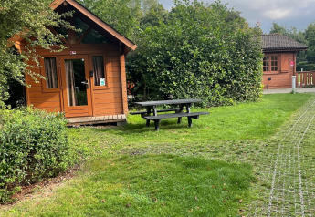 Foto einer Basic Log Cabin bei Urban-Gardens Ieper, Belgien, mit grünem Rasen und Sitzbank im Vordergrund.