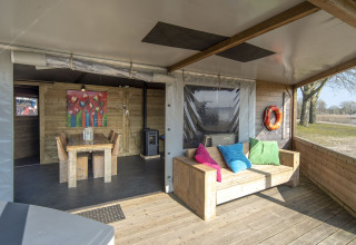 Covered patio of Dune house at Marina Parcs Almere, Netherlands, featuring wooden furniture and bright pillows.