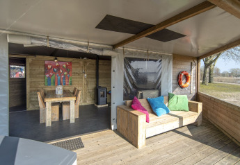Covered patio of Dune house at Marina Parcs Almere, Netherlands, featuring wooden furniture and bright pillows.