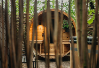 Blick durch Bäume auf ein rundes Holzhaus bei Boshuys Ultra Wellness, Buitengoed Ruysbos, Niederlande.