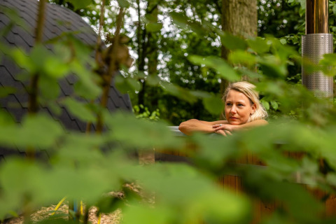 Woman relaxing in a wooden hot tub surrounded by greenery at Boshuys Ultra Wellness, Buitengoed Ruysbos, Netherlands.